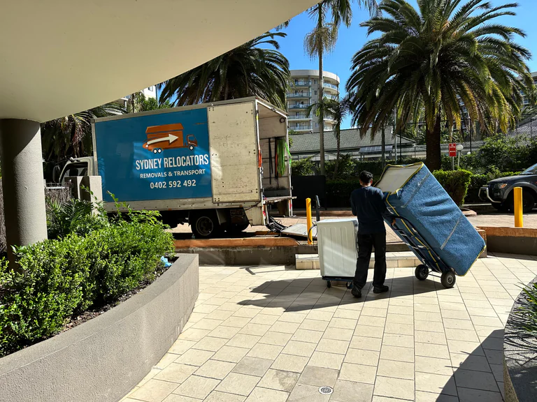 A washing machine and a wrapped mattress trolleyed across an apartment forecourt